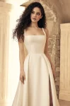 A woman stands in the ivory P5150 by Paloma Blanca gown, featuring a sleeveless, square-neck design with a high slit, paired with strappy heels, set against classical architectural elements and neutral tones.