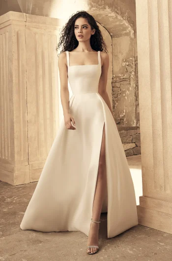 A woman stands in the ivory P5150 by Paloma Blanca gown, featuring a sleeveless, square-neck design with a high slit, paired with strappy heels, set against classical architectural elements and neutral tones.