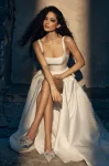 A woman stands in the ivory P5150 by Paloma Blanca gown, featuring a sleeveless, square-neck design with a high slit, paired with strappy heels, set against classical architectural elements and neutral tones.
