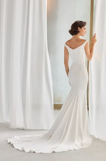 A woman in an off-the-shoulder white P5211 by Paloma Blanca wedding gown, featuring a fitted bodice and long train, stands facing a window and touches a curtain.