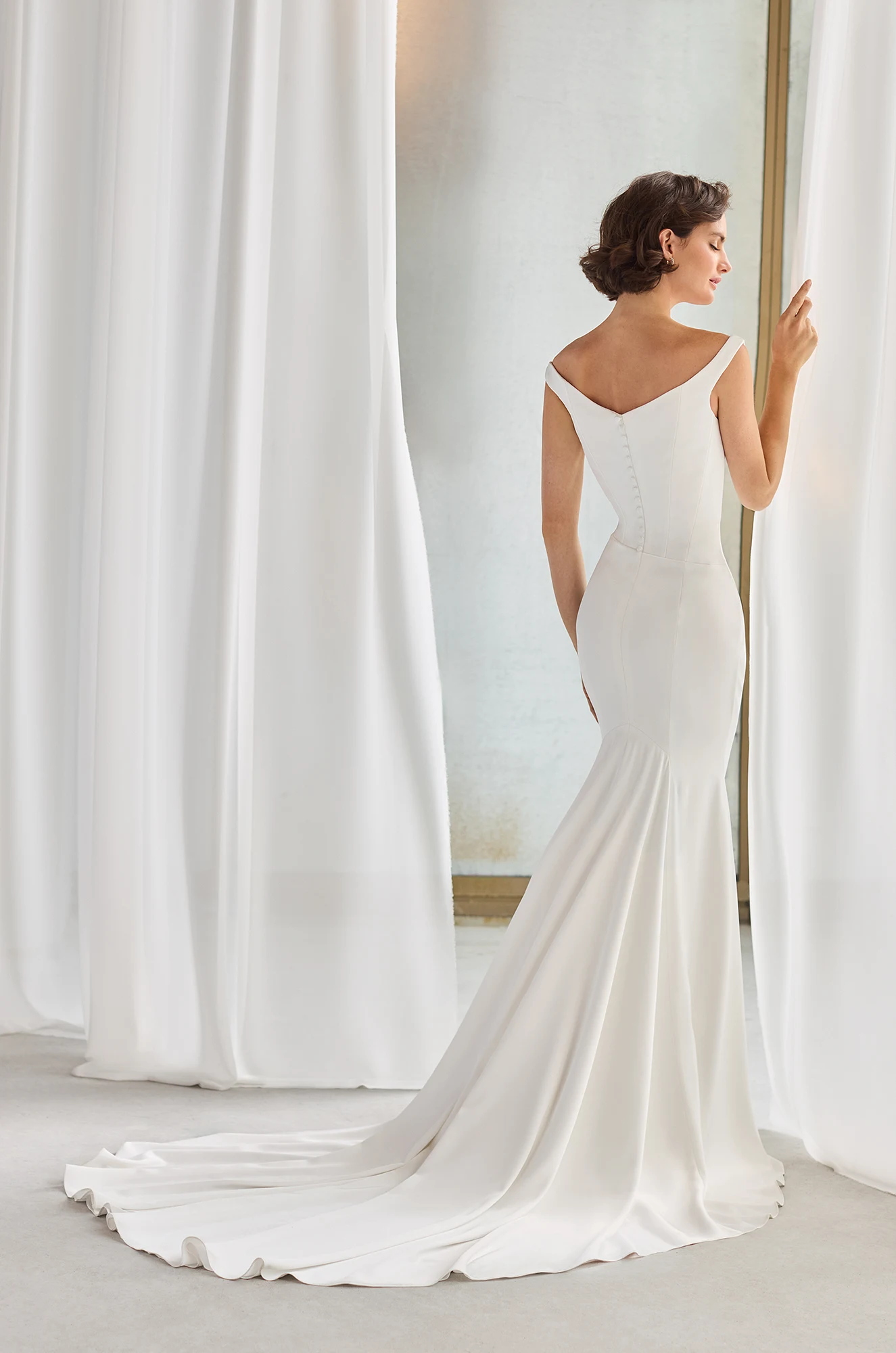 A woman in an off-the-shoulder white P5211 by Paloma Blanca wedding gown, featuring a fitted bodice and long train, stands facing a window and touches a curtain. A woman in an off-the-shoulder white P5211 by Paloma Blanca wedding gown, featuring a fitted bodice and long train, stands facing a window and touches a curtain.
