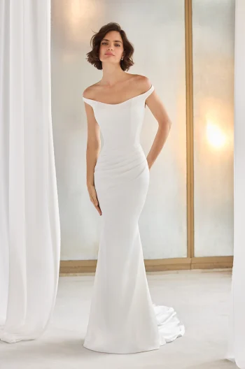 A woman stands indoors near white curtains and a frosted window, wearing the P5211 by Paloma Blanca—an off-the-shoulder, fitted white gown with a slight train.