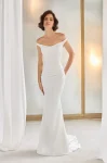 A woman stands indoors near white curtains and a frosted window, wearing the P5211 by Paloma Blanca—an off-the-shoulder, fitted white gown with a slight train.